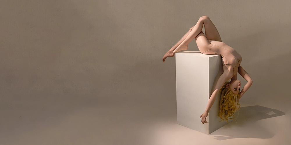 Person with long blond hair draped upside down over a white pedestal against a neutral beige background.