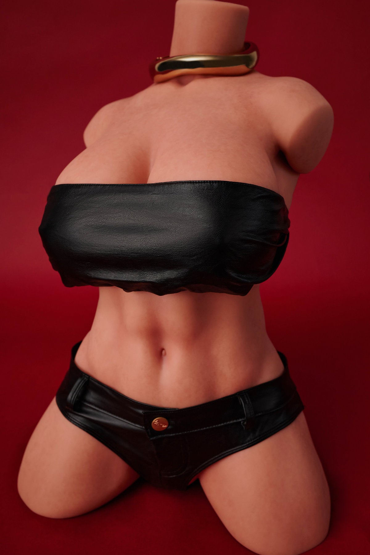 A frontal view of the headless CLIMAX DOLL - ULW 663# Silicone Torso (Black) wearing a gold collar necklace, a black leather-look tube top, and matching low-rise black shorts against a red background.