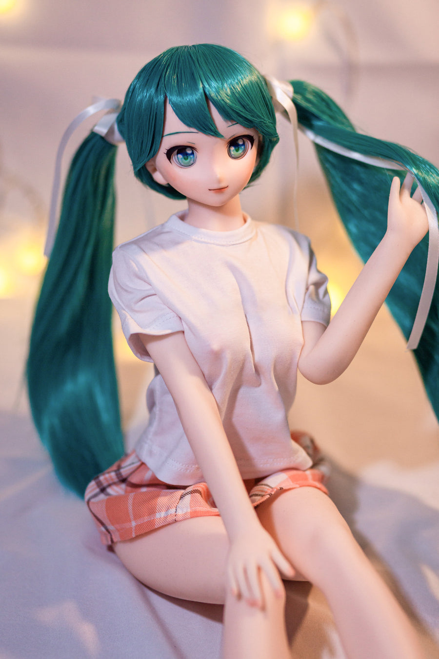 CLIMAX DOLL Mini J60 cm Hybrid - Miku (Cinnamon) - Seated on soft white bedding with one hand lifting a long teal twin tail, white tee and pink plaid mini skirt, warm fairy-light bokeh behind.