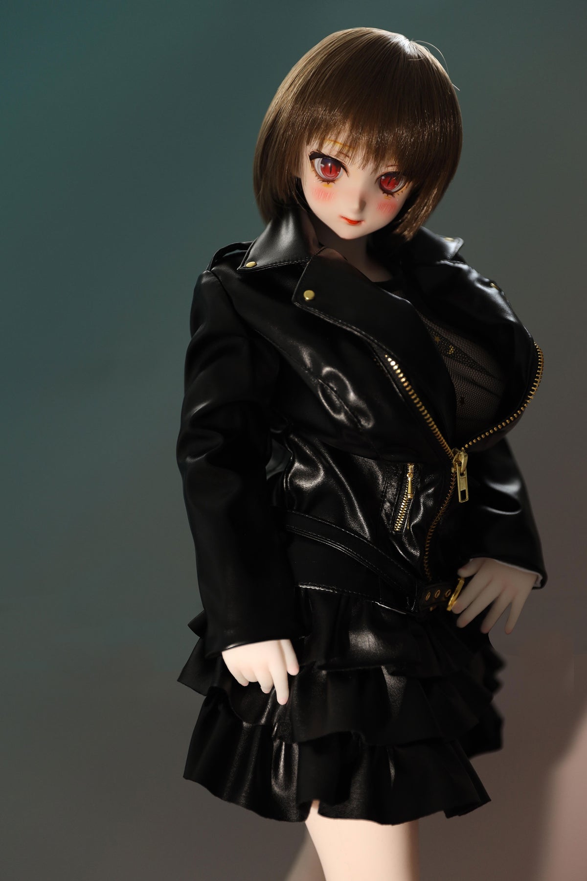 CLIMAX DOLL Mini J60cm P Hybrid - Zella (White) - Close-up of the glossy black ruffled jacket-dress, showing the gold zipper and tiered ruffle hem, short brown bob and red-brown eyes, teal-green backdrop.