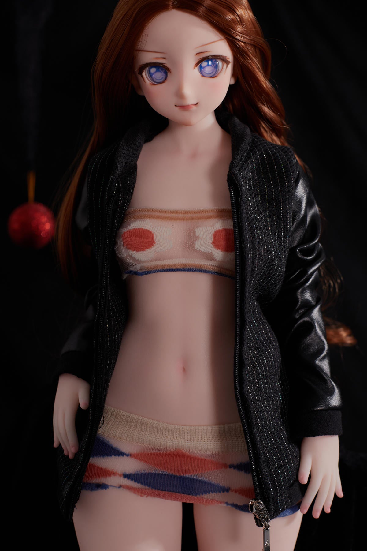 CLIMAX DOLL Mini J60cm XS Hybrid - Nanami (Cinnamon) standing pose in an open black knit cardigan, patterned tube top, and colorful geometric mini skirt, long auburn hair, violet anime eyes, black backdrop with red ornament accents