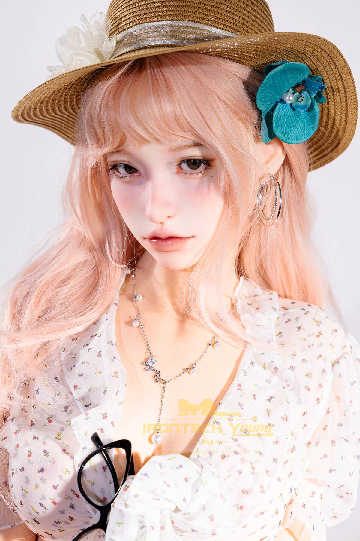 Irontech Doll 166 cm 2.0 Young Series E Silicone - Mio close-up portrait, straw sun hat with a teal flower accent, wavy pale pink hair, white floral dress, layered necklaces, sunglasses hanging from her collar, bright white studio backdrop
