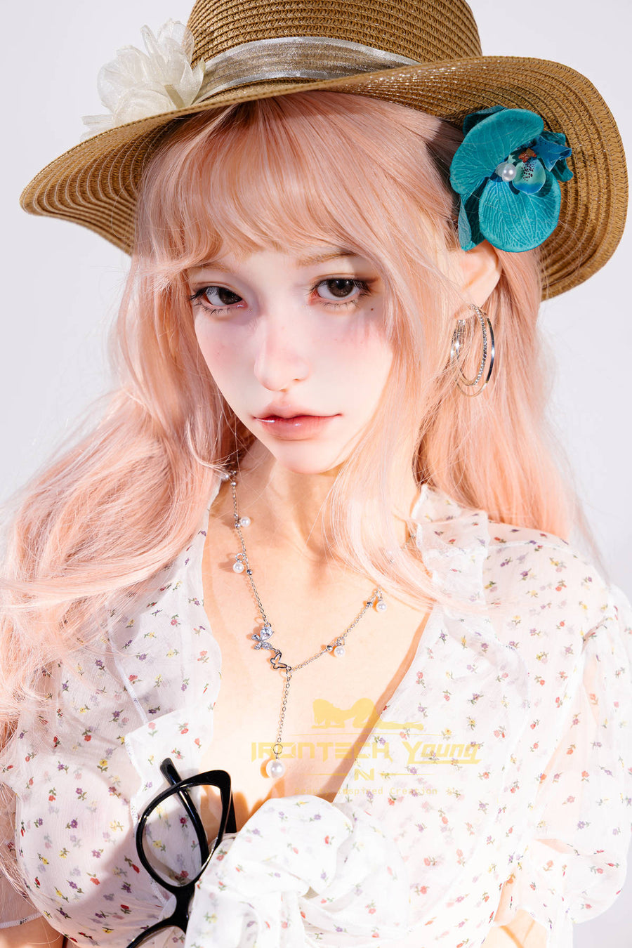 Irontech Doll 166 cm 2.0 Young Series E Silicone - Mio close-up portrait, straw sun hat with a teal flower accent, wavy pale pink hair, white floral dress, layered necklaces, sunglasses hanging from her collar, bright white studio backdrop