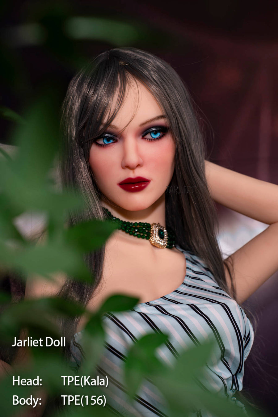 Jarliet Doll 156 cm B TPE - Kala V2 close-up portrait, dark straight hair with bangs, bold blue eyeshadow, red lips, black choker with pendant, black-and-white striped playsuit, hand raised near her hair, green foliage in the foreground, warm stadium lights in the background