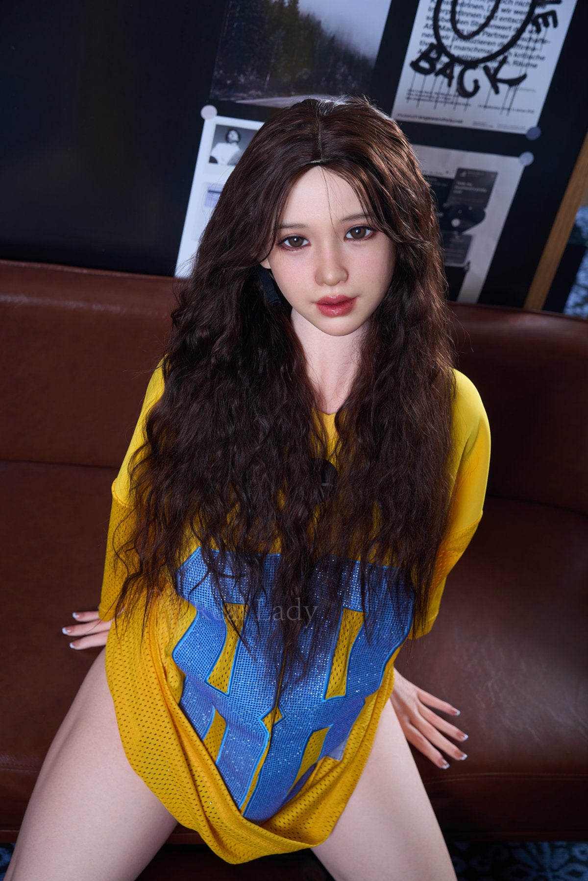 RealLady 159cm H Silicone - Alodia V3 – Seated pose, mustard-yellow off-shoulder dress, long wavy brown hair, dark interior backdrop.