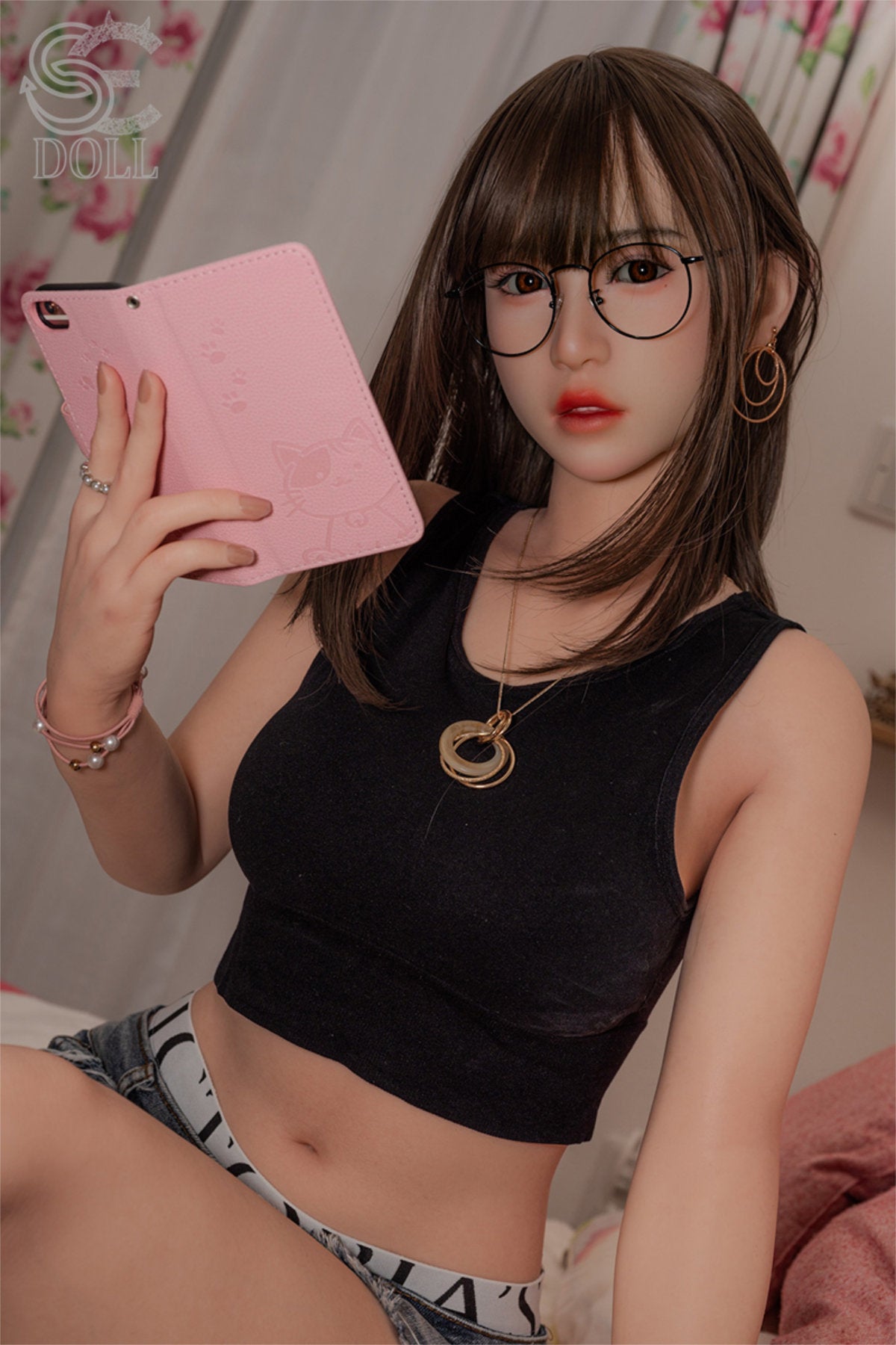 SEDOLL T153cm A Aya Series Silicone - Sena.A – Seated pose holding a pink book prop, black cropped tank top with denim skirt, round eyeglasses, dark brown bangs and long hair, cozy bedroom backdrop with floral wallpaper.