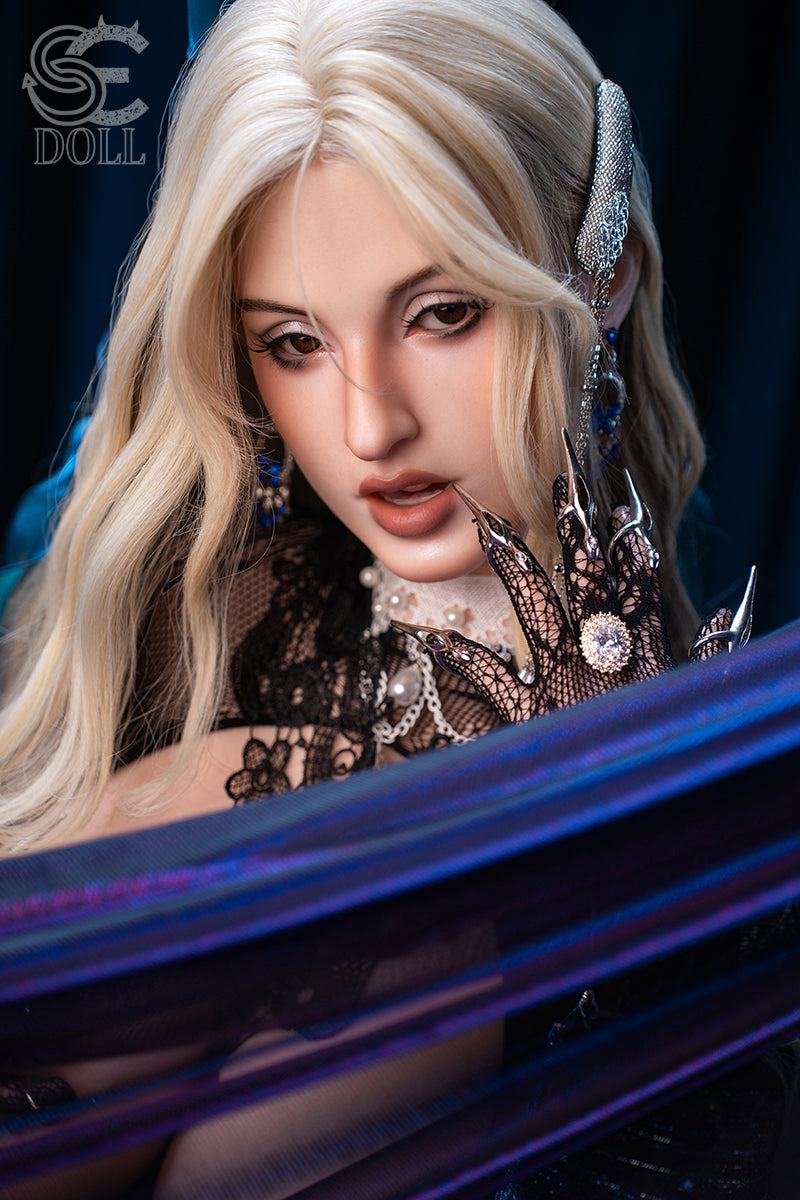 SEDOLL T155 cm J Silicone Pro - Cleopatra.A close-up portrait, wavy platinum blonde hair, an elaborate metallic chain headdress draping down one side of her face, blue satin fabric over one shoulder, moody dark navy backdrop