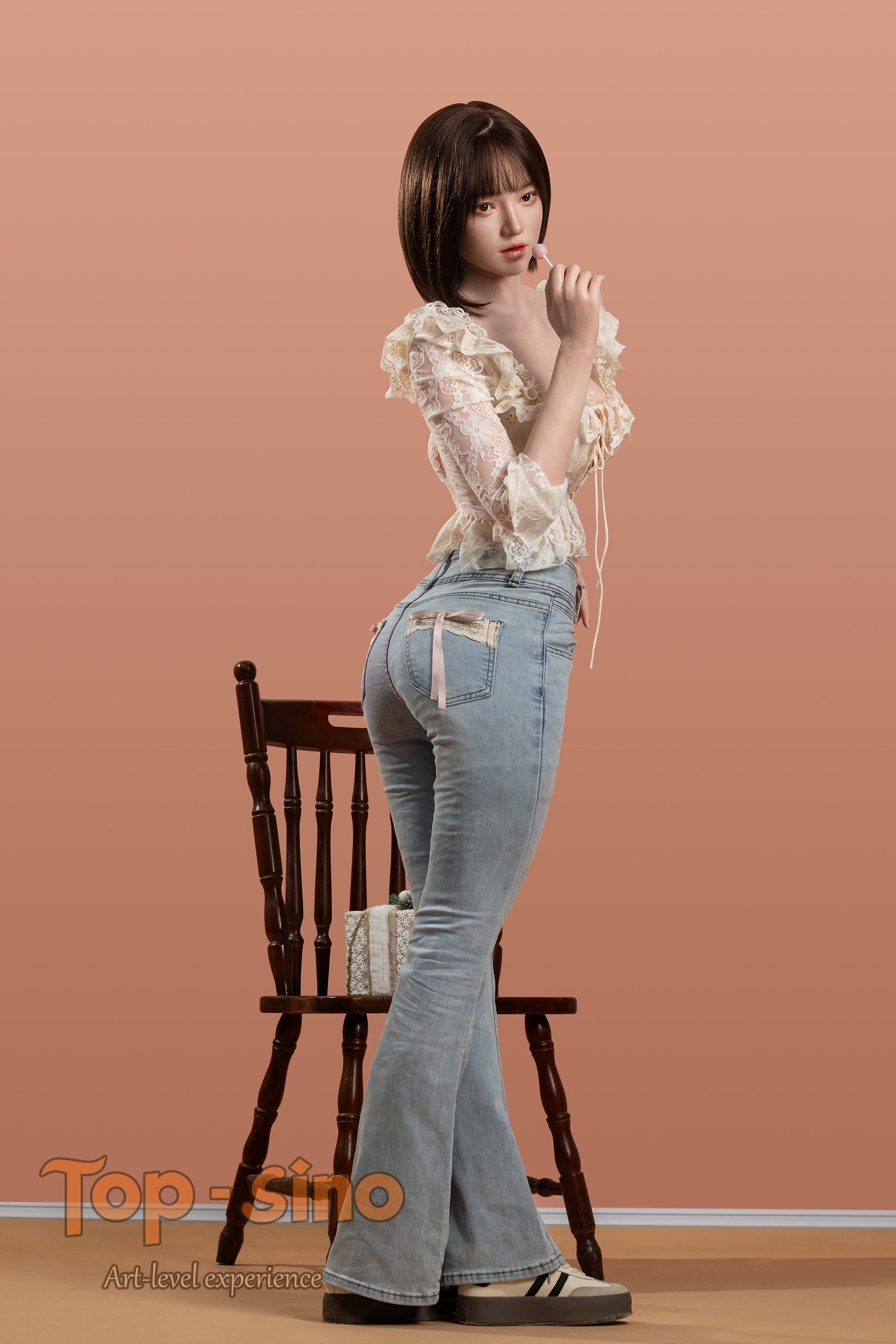 Top Sino 167 cm Platinum Silicone - Mimu (RRS+) - V2 in a white blouse and blue jeans standing in front of a wooden chair against a peach wall.